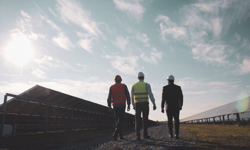 Three mixed ethnic men in uniform and hard hat walking around solar power station and examining it. Side view