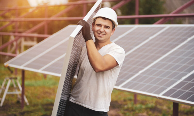 Smiling young worker in protective helmet carrying big shiny solar photo voltaic panel on exterior metal platform background on sunny summer warm day. Renewable ecological green energy concept.